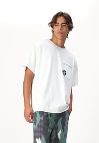 T-shirt blanc surdimensionné avec une poche poitrine et des détails de couture noire, associé à un pantalon à motif argyle multicolore.
