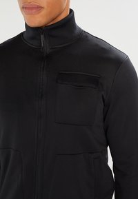 Schwarze Zip-Jacke mit hohem Kragen, strukturiertem Stoff und einer Brusttasche mit Klappe. Sie verfügt über zwei Fronttaschen zur Aufbewahrung.