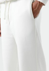 Pantalon de jogging en coton blanc avec poches latérales, présentant une coupe décontractée et une taille élastique avec cordon de serrage. Comprend des coutures décoratives discrètes.