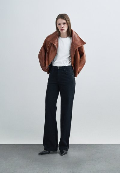 Καφέ δερμάτινο μπουφάν με φαρδιές πέτσες, λευκό μπλουζάκι και μαύρο παντελόνι με φαρδιά μπατζάκια. Κομψά μαύρα παπούτσια με μυτερή μύτη ολοκληρώνουν την εμφάνιση.