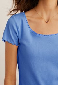 Top blu ribbed con scollatura a barca, orli delle maniche a scallop e rifiniture a contrasto. Collana dorata con un pendant rotondo viola.