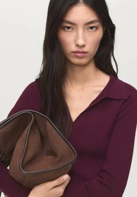 Sac en tissu marron avec des bordures noires. Modèle portant un haut ajusté bordeaux foncé avec un col. Cheveux noirs raides. Fond neutre.