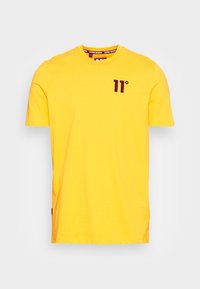 Żółta koszulka z krótkim rękawem z purpurowym logo "11°" na lewej piersi oraz dopasowanym purpurowym napisem na wewnętrznej części kołnierza.