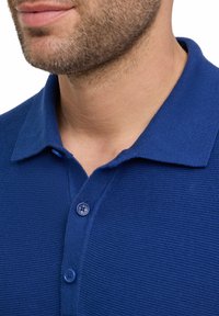 Niebieska koszulka polo wykonana z teksturowanej tkaniny, z kołnierzem i zapięciem na trzy guziki. Posiada okrągłe guziki i lekką konstrukcję.