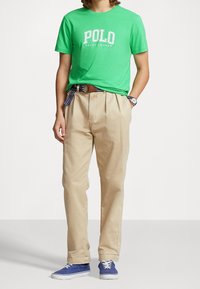 Zelené bavlněné tričko s logem "POLO", béžové skládací chinos kalhoty, modré tenisky a vícebarevný pruhovaný pásek.