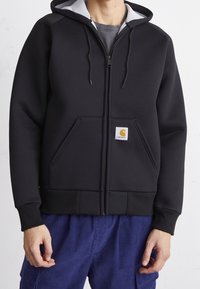 Sudadera con cremallera en negro, con forro de capucha gris, dos bolsillos frontales y un pequeño logotipo cuadrado. Fabricada en un tejido suave y grueso. Puños y dobladillo acanalados.