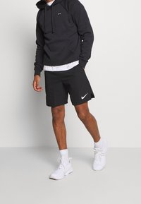 Uomo che indossa un hoodie nero di Under Armour, pantaloni corti neri di Nike, calze bianche e sneaker bianche, in piedi contro uno sfondo chiaro e semplice.