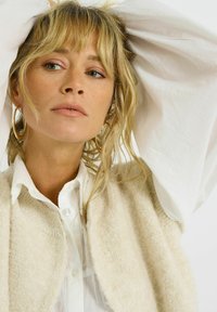 Mujer con cabello rubio y ojos verdes, vistiendo grandes pendientes de aro, una camisa blanca y un chaleco texturizado beige, contra un fondo sencillo.