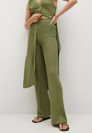 Frau trägt olivgrüne, texturierte, gestrickte, weite Hose und passende ärmellose lange Weste mit Knöpfen sowie schwarze Sandalen.