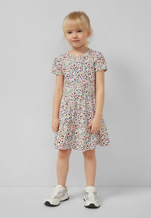 Jeune fille debout en robe blanche à motifs floraux et baskets blanches sur fond gris uni.