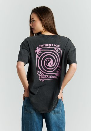 Vrouw met lang bruin haar, gekleed in een zwart oversized T-shirt met een roze spiraal en de tekst "Saltwater Soul Balanced Roll", gecombineerd met een blauwe spijkerbroek, handen in de zakken.