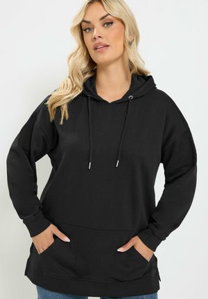 Blond kvinde iført en sort hættetrøje med lommer, stående med hænderne i lommerne mod en ensfarvet lys baggrund.