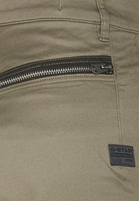Khaki kalhoty s kapsou na zip, texturovanou tkaninou a černou značkovou etiketou s nápisem "G-Star RAW" na postranním švu.