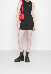 Robe sans manches noire au design ajusté, associée à un sac bandoulière rouge et des bottes de combat noires à lacets avec une semelle épaisse.