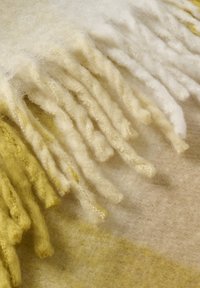 Plaid di lana con bordi frangiati, con una sfumatura che va dal bianco a toni di giallo pallido, che mostra una superficie testurizzata e morbida.