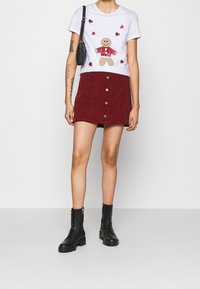 Bílý tričko s grafikou perníkového mužíka a červenými srdíčky, doplněné burgundskou manšestrovou sukní se metalovými knoflíky. Černé šněrovací kotníkové boty.