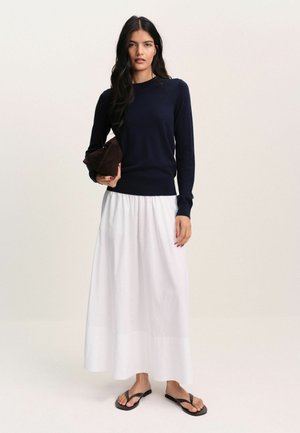 Frau trägt ein marineblaues Langarmtop, weißen knöchellangen Rock, schwarze Flip-Flops und hält eine kleine dunkelbraune Clutch.