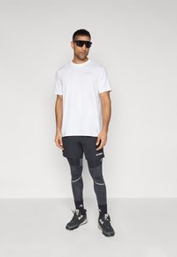 Camiseta blanca de manga corta, pantalones cortos negros, leggings con estampado, zapatillas negras y gafas de sol; presenta acentos de marca en la ropa.