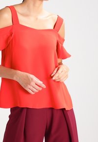 Červený top bez ramínek se čtvercovým výstřihem a volánkovými detaily, vyrobený z lehkého materiálu, v kombinaci s burgundskými kalhotami širokého střihu.