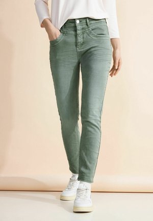 Femme portant un jean vert slim, un haut à manches longues blanc et des baskets blanches, avec une main dans la poche, debout sur un fond neutre.