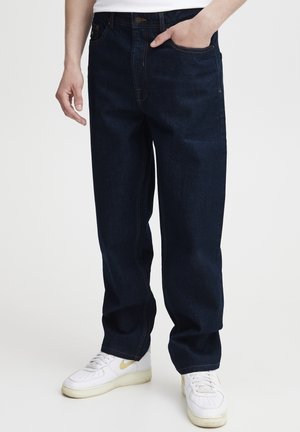 Jeans de mezclilla azul oscuro con un corte relajado, con costuras naranjas y bolsillos frontales. Combinados con zapatillas blancas.