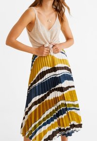 Falda plisada en amarillo, azul, marrón y franjas blancas; combinada con una camiseta sin mangas beige claro con un lazo en el frente y detalles de botones.