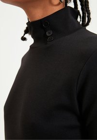 Haut noir à col montant en tissu texturé, doté d'une patte de boutonnage avec trois boutons assortis sur le côté.