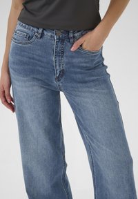 Donna che indossa jeans blu medio a gamba dritta con la mano in tasca e una maglietta scura su uno sfondo neutro.