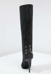 Bota de ante negra hasta la rodilla con punta afilada, tacón de aguja y detalles de cordones en la parte posterior. Textura suave y diseño elegante.