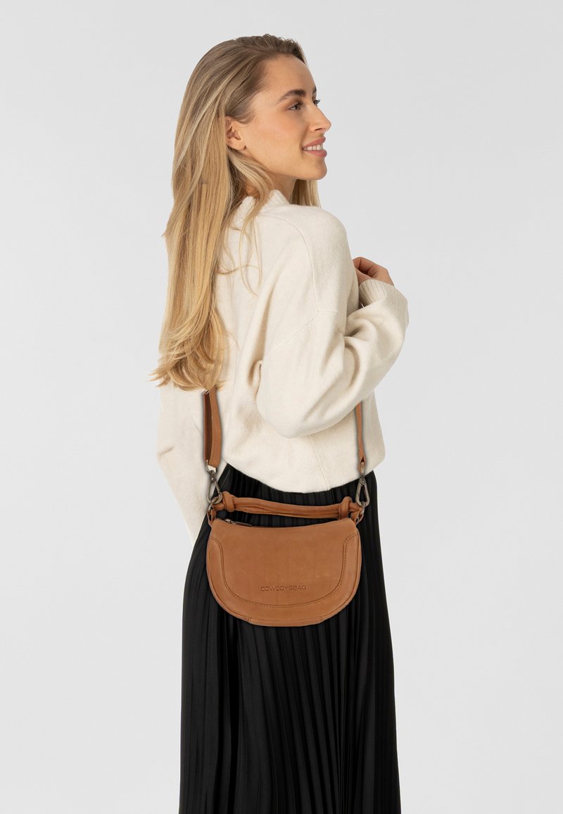 Tan leren crossbody tas met een gebogen vorm, verstelbare schouderband en logo-detail. Model draagt een crèmekleurige trui en een zwarte geplooide rok.