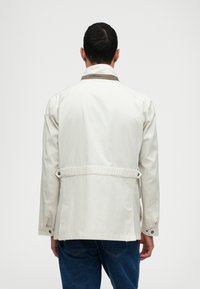 Chaqueta de color beige claro con un cuello marrón. Presenta una cintura ajustada, puños con botones y una textura suave. Vista desde la espalda, combinada con vaqueros azules.