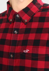 Czerwona i czarna koszula w kratkę z guzikiem, kołnierz, cztery widoczne guziki oraz biała wyszyta logo ptaka na kieszeni piersiowej.