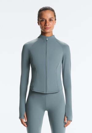 Frau trägt eine enganliegende, langärmlige, hellgraue Jacke mit Reißverschluss und passende Leggings, steht vor einem schlichten weißen Hintergrund.