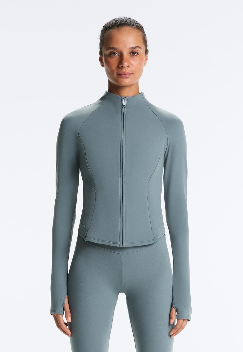 Femme portant une veste zippée ajustée à manches longues gris clair et un legging assorti, debout devant un fond blanc uni.