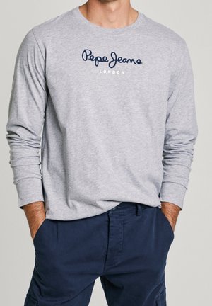 Mężczyzna w jasnoszarej koszuli z długim rękawem z napisem "Pepe Jeans London" i granatowych spodniach, ręce w kieszeniach.