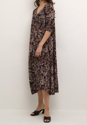 Kvinde stående på siden iført en løs, mønstret midikjole med halvlange ærmer og sorte åbne hæl-sandaler mod en ensfarvet baggrund.