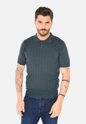 Uomo con capelli corti e scuri e barba, che indossa una polo a maniche corte verde scuro e jeans blu, con tatuaggi visibili su entrambe le braccia.