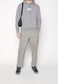 Sweat à capuche gris avec poche avant et logo, associé à un pantalon à carreaux larges noir et blanc, et des baskets bleues. Sac à bandoulière noir.