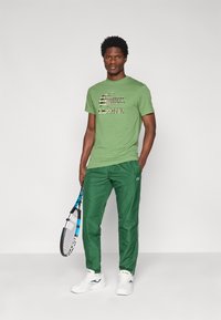 Camiseta de algodón verde con un patrón gráfico, combinada con pantalones deportivos verdes. Zapatillas deportivas blancas y una raqueta de tenis completan el conjunto.