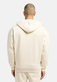 Beige hætte sweatshirt med en blød tekstur, ribbede manchetter og kant, samt et præget logodetalje nær bunden.