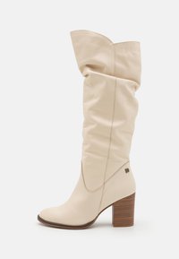 Botas de cuero beige hasta la rodilla con un diseño holgado, puntera afilada y un tacón marrón apilado. Presenta costuras sutiles y un detalle de marca.