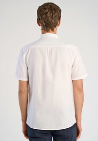 Camisa de manga corta de color claro fabricada en lino. Presenta un collar, puños enrollados y un dobladillo ligeramente curvado en la parte trasera.