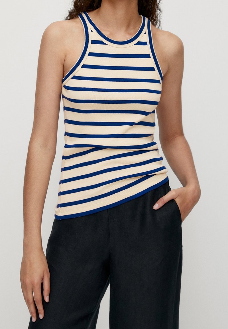 Top sans manches rayé en crème et bleu, avec un col rond et une silhouette ajustée. Porté avec un pantalon sombre, texture lisse.