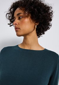 Dunkles Petrol, gerippter Pullover mit rundem Halsausschnitt. Lockiges Haar, das das Gesicht umrahmt, mit einem goldenen Creolenohrring als Akzent.