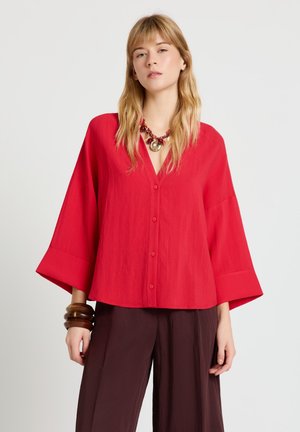 Frau trägt eine locker sitzende rote Bluse mit Knöpfen und weiten Ärmeln, dunkelbraune Hose, klobige hölzerne Armbänder und eine rote auffällige Halskette vor hellem Hintergrund.