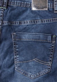 Achterzak van denim jeans met contrasterende witte stiksels. Materiaal is blauwe denim, met een gestructureerd oppervlak en een rechthoekige vorm.