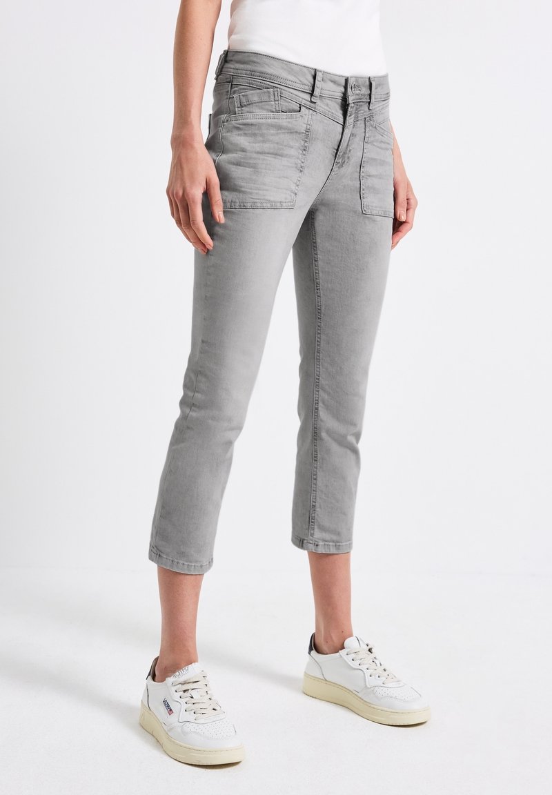 Personne portant un jean gris court et des baskets blanches, debout devant un fond blanc uni.