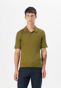 Polo de punto verde con mangas cortas, que presenta una textura acanalada y cuello. Combinado con pantalones oscuros, resaltando un diseño ajustado.