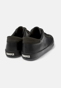 Adidași negri din piele cu o textură netedă, având o talpă din cauciuc gri și un guler căptușit. Numele brandului "CAMPER" este vizibil pe călcâi.