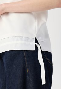 Hvit sportstopp med lagdelt nederkant og bånddetalj på siden, kombinert med mørk denimjeans med gul søm.